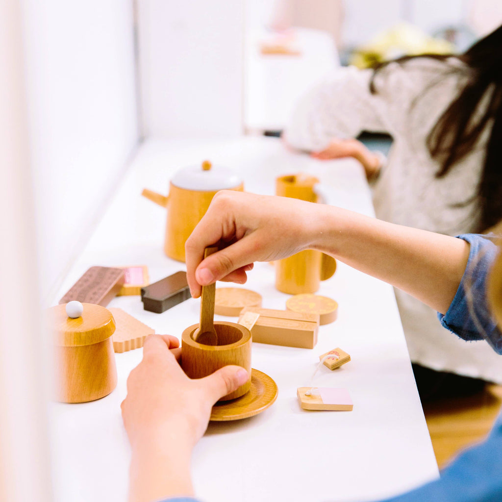 A child indulges in imaginative play with the Wooden Tea Set on a white surface, featuring cups, teapots, and food pieces. Another child joins the fun in this charming scene reminiscent of designer coffee table books.