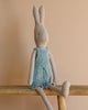 From the enchanting bunny & rabbit collection, the Maileg Rabbit size 4 in a blue knitted overall sits cozily on a wooden bench against a soft beige backdrop.