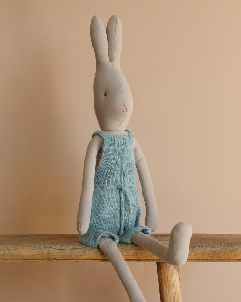 From the enchanting bunny & rabbit collection, the Maileg Rabbit size 4 in a blue knitted overall sits cozily on a wooden bench against a soft beige backdrop.
