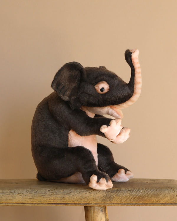 The Elephant Calf Sitting Stuffed Animal from HANSA, with realistic and hand-sewn details, sits on a wooden stool against a beige background. Its raised trunk and bent arm give it a playful charm in brown and beige.