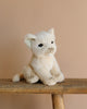 The Lion Cub Sitting Stuffed Animal, with realistic features, light beige fur, and dark eyes, sits on a wooden stool against a plain beige background.