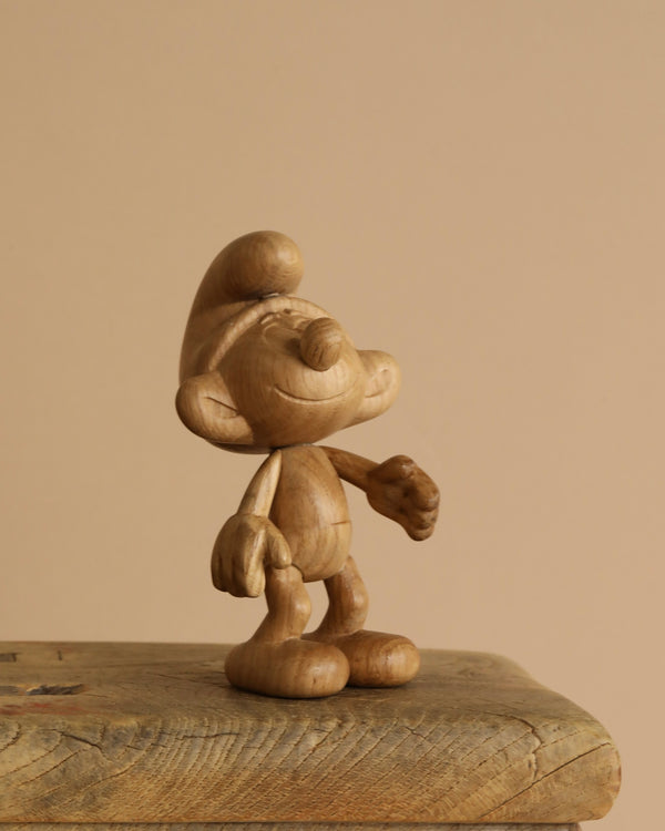 An "Oak Smurf" wooden sculpture stands on an aged wooden surface, evoking a sense of adventure in Smurf Village. The figure features a round nose, large ears, and a hat, set against a simple beige background.