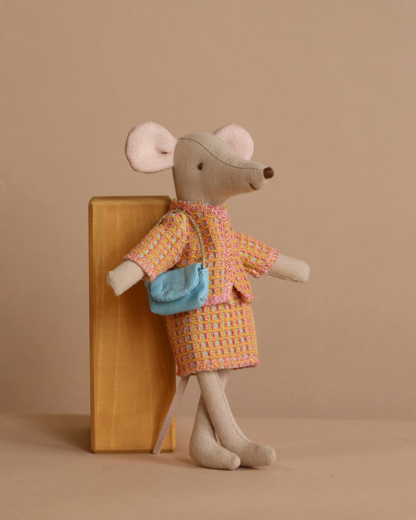 The Maileg Mom Mouse With Extra Outfit - Petite Couture stands against a wooden block in a vibrant checkered coat, clutching a tiny blue purse. A simple beige backdrop highlights the charm of this delightful plush toy's outfit.