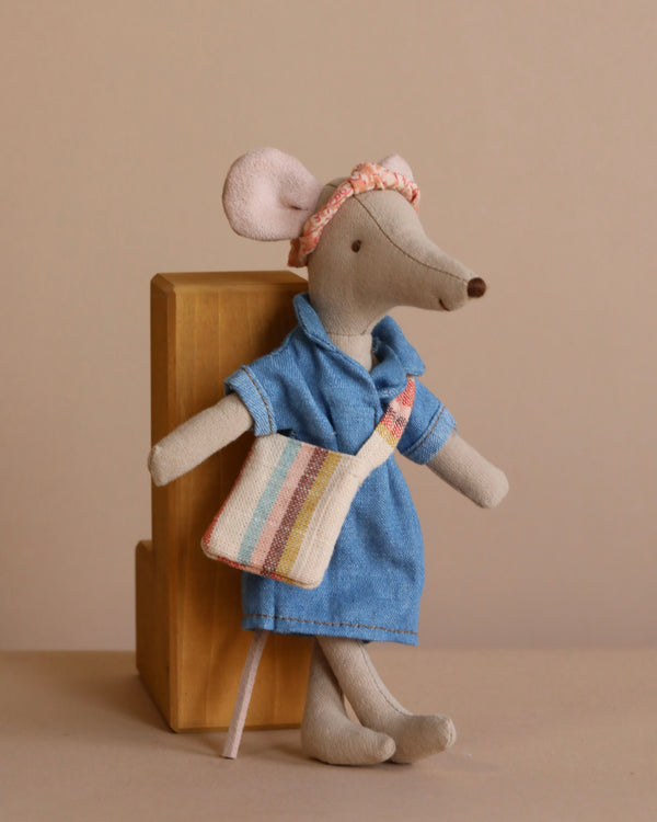 The Maileg Mom Mouse With Extra Outfit - Petite Couture, wearing a denim-like blue dress, pink headband, and striped bag, leans against a wooden block. This cotton-crafted toy adds warmth to the neutral beige background.