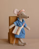 The Maileg Mom Mouse With Extra Outfit - Petite Couture, wearing a denim-like blue dress, pink headband, and striped bag, leans against a wooden block. This cotton-crafted toy adds warmth to the neutral beige background.