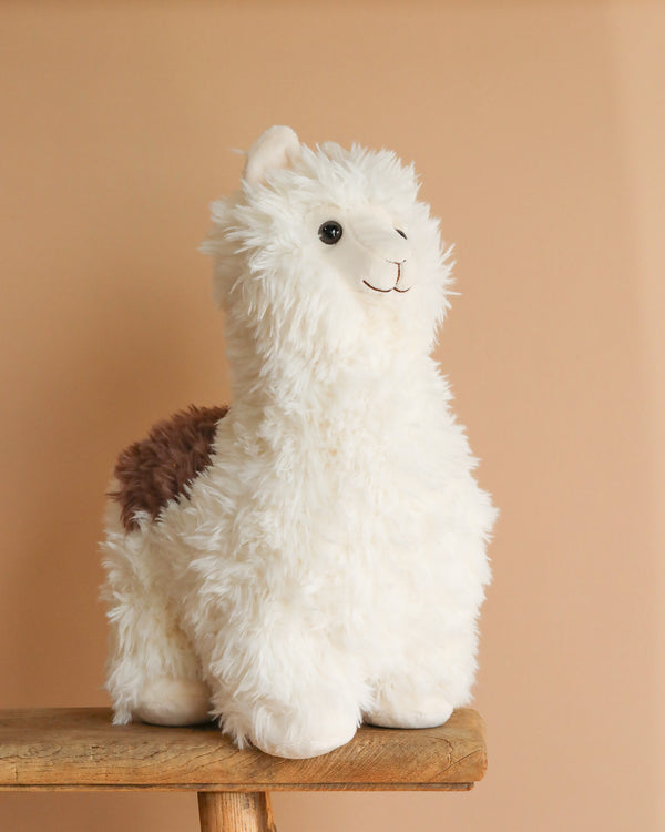 The Snuggleluvs Alpaca Weighted Stuffed Animal 15", featuring a fluffy white body with a brown patch, rests serenely on a wooden stool against a beige background, capturing the charm of Wild Republic's plush toys.