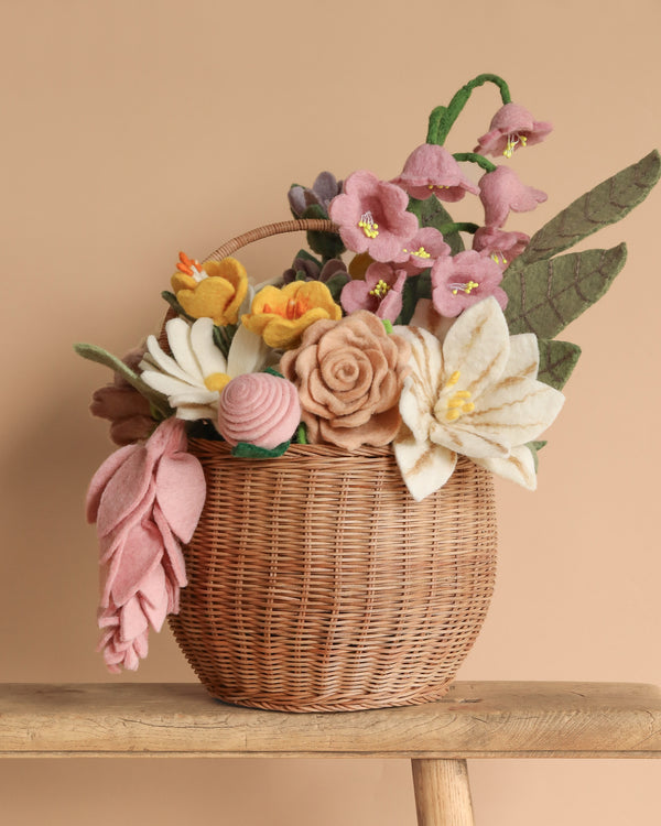 The Basket Of Flower Flowers, a wicker basket brimming with pastel eco-friendly felt blooms like lilies and roses, rests on a wooden surface. This felt flower arrangement features handcrafted flowers in various textures and muted tones against a beige background.