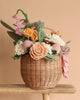 The "Basket Of Flower Flowers" showcases a woven basket filled with eco-friendly felt flowers, including handcrafted roses and lilies in pastel pink, yellow, white, and purple. It sits on a wooden surface against a beige backdrop.