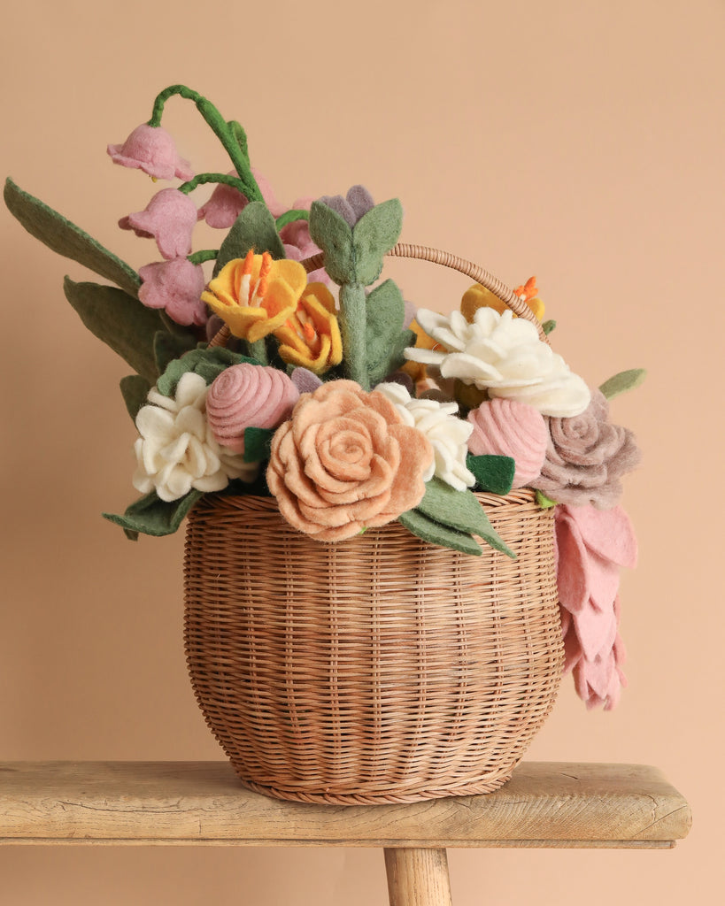 The "Basket Of Flower Flowers" showcases a woven basket filled with eco-friendly felt flowers, including handcrafted roses and lilies in pastel pink, yellow, white, and purple. It sits on a wooden surface against a beige backdrop.