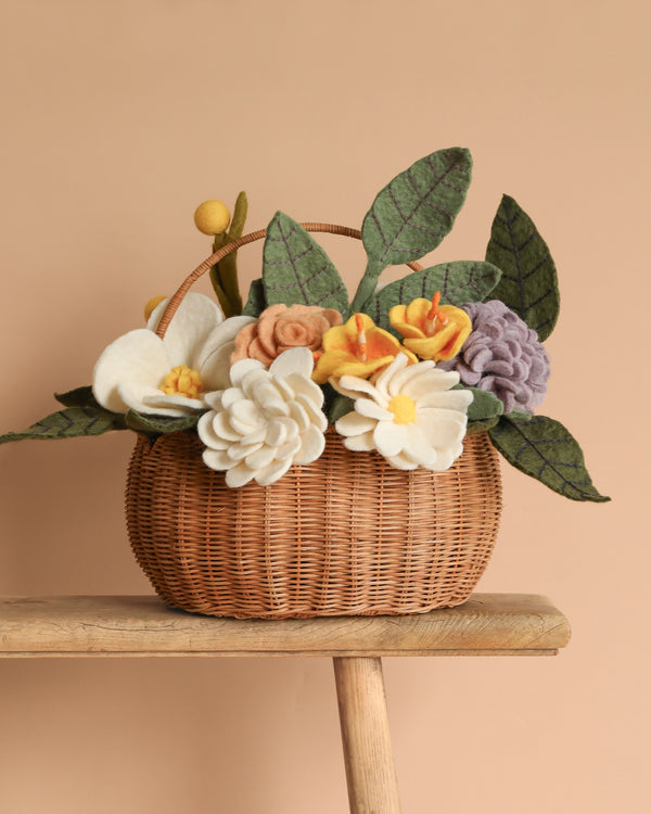 The Basket Of Felt Flowers, filled with eco-friendly handcrafted blooms in cream, peach, yellow, and lavender hues, sits on a wooden stool against a warm beige background.