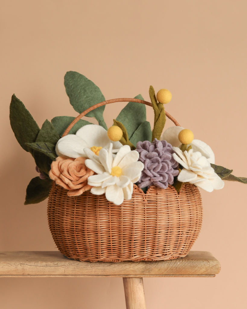 The Basket of Felt Flowers, handcrafted and eco-friendly, features a wicker basket with soft white, peach, and purple felt flowers surrounded by green leaves on a wooden surface against a neutral beige background.