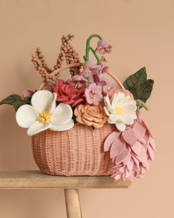 The Basket of Felt Flowers, featuring eco-friendly handcrafted blooms in white, pink, and orange with lush green leaves, is elegantly displayed on a wooden surface against a calming beige background.