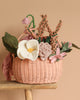 The "Basket of Felt Flowers" features a woven design adorned with handcrafted blooms and felt flowers, including a large white bloom, pink rose, green leaves, and small pink buds. This eco-friendly piece sits on a wooden surface against a beige background.
