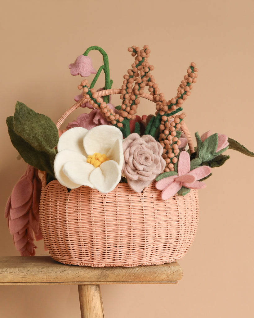 The "Basket of Felt Flowers" features a woven design adorned with handcrafted blooms and felt flowers, including a large white bloom, pink rose, green leaves, and small pink buds. This eco-friendly piece sits on a wooden surface against a beige background.