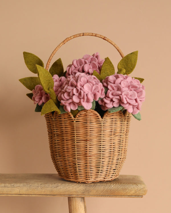 The Basket Of Felt Flowers - Hydrangea, set in a wicker basket and featuring pink felt flowers with green felt leaves, is elegantly displayed on a wooden plank against a beige background.