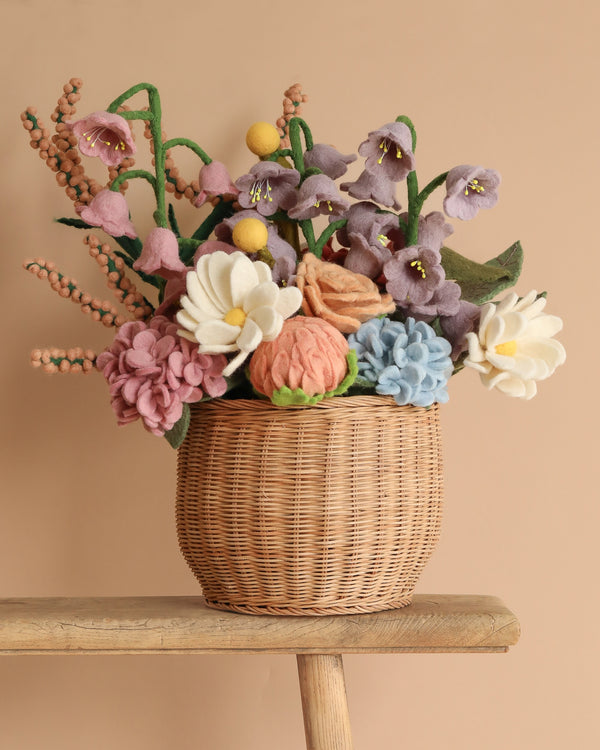 The "Basket Of Felt Flowers" features eco-friendly, non-toxic felt blooms in pink, purple, blue, and white. Set in a wicker basket on a wooden surface with a beige background, these handcrafted pieces showcase detailed designs that beautifully mimic real flowers.