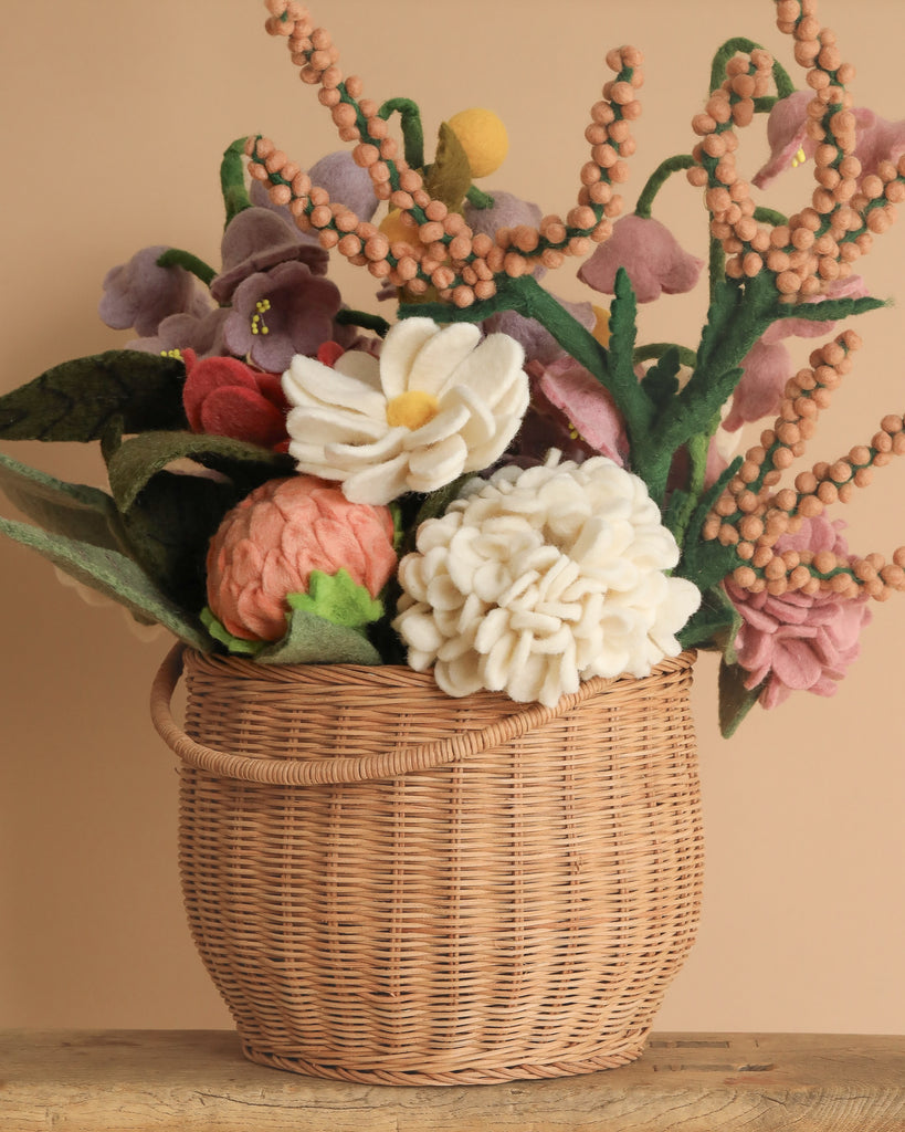 The Basket Of Felt Flowers is a woven basket filled with eco-friendly felt blooms, including daisies and roses. These vibrant, handcrafted flowers are artfully arranged in an organic style against a neutral background.