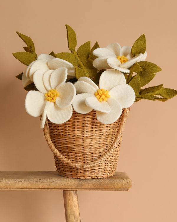 The Basket Of Felt Flowers - Magnolia, featuring handcrafted felt magnolias and green leaves, adds a stunning touch of sustainable home decor when displayed on a wooden stool against a beige background.