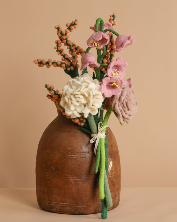 The Handcrafted Felt Flower Bouquet includes pink blossoms, a large white bloom, green stems with orange berry-like accents, all arranged in a rustic brown ceramic vase against a light brown backdrop.