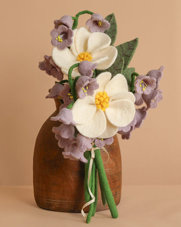The Handcrafted Felt Flower Bouquet includes white and yellow flowers with purple bell-shaped blossoms, displayed in a rustic brown vase on a neutral background. Two green felt leaves beautifully contrast the sustainable floral design.