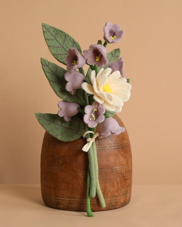 The Handcrafted Felt Flower Bouquet features sustainable felt florals, including green leaves, small purple blossoms, and a large white flower with a yellow center, arranged in a wooden vase against a beige background.