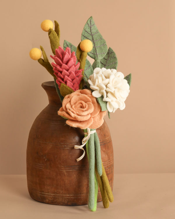 The Handcrafted Felt Flower Bouquet features wool-felt blooms in pink, peach, and white with green and yellow accents in a rustic brown vase against a soft beige backdrop.