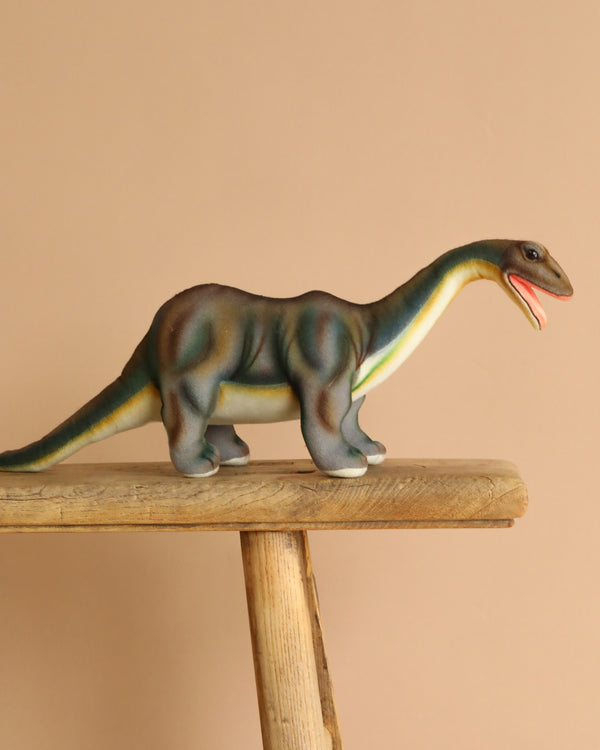 A Brontosaurus Dinosaur Stuffed Animal with a long neck and greenish-brown skin stands on a wooden surface against a beige background. Its open mouth, revealing pink hues inside, highlights realistic features that capture the creature's ancient majesty.