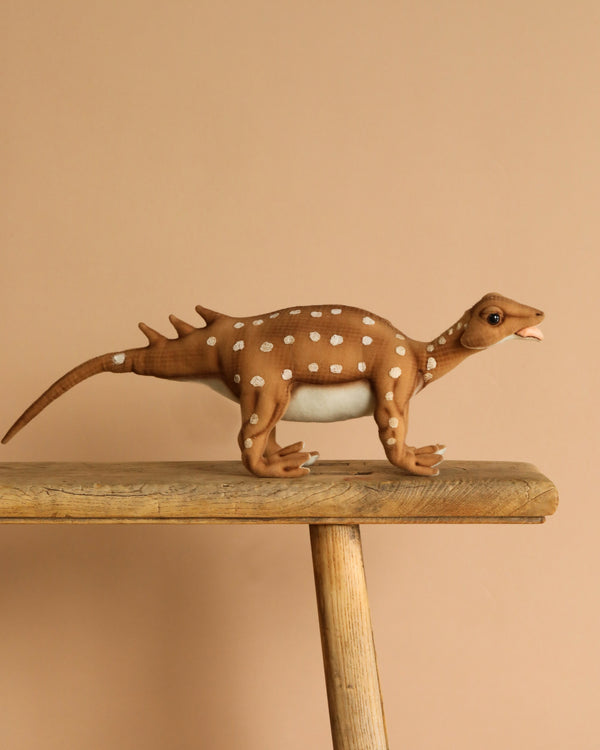 The Minmi Paravertebra Dinosaur Stuffed Animal stands on a wooden surface with a beige background. It features a brown body with white spots, boasting realistic details. Its raised tail and slightly open mouth give it unique charm.