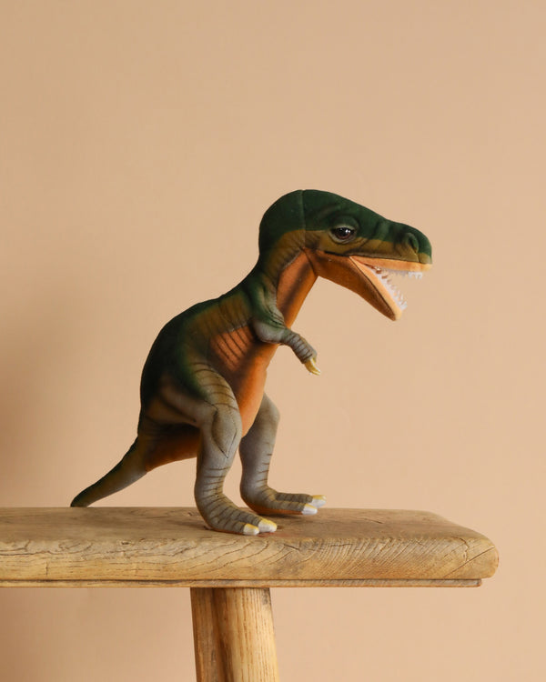 A plush T-Rex Dinosaur Stuffed Animal with a green body and lighter underbelly stands upright on a wooden stool against a beige background, featuring realistic details including an open mouth showing its teeth.