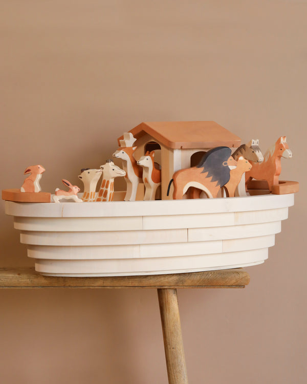 The Noah's Ark With Animals toy set, with its intricately painted lions and giraffes on a stylized boat and small house, captures the essence of Holztiger Figures. Made in Europe, it sits on a wooden surface against a warm beige backdrop.