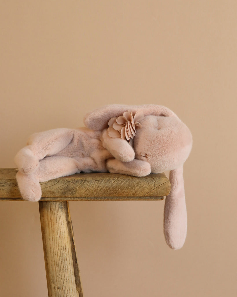 A soft Maileg Sleeping Bunny With Bed, crafted from recycled polyester, lies on its side on a rustic wooden shelf. The light pink plush toy features floppy ears and a decorative fabric flower near its head against a beige background.