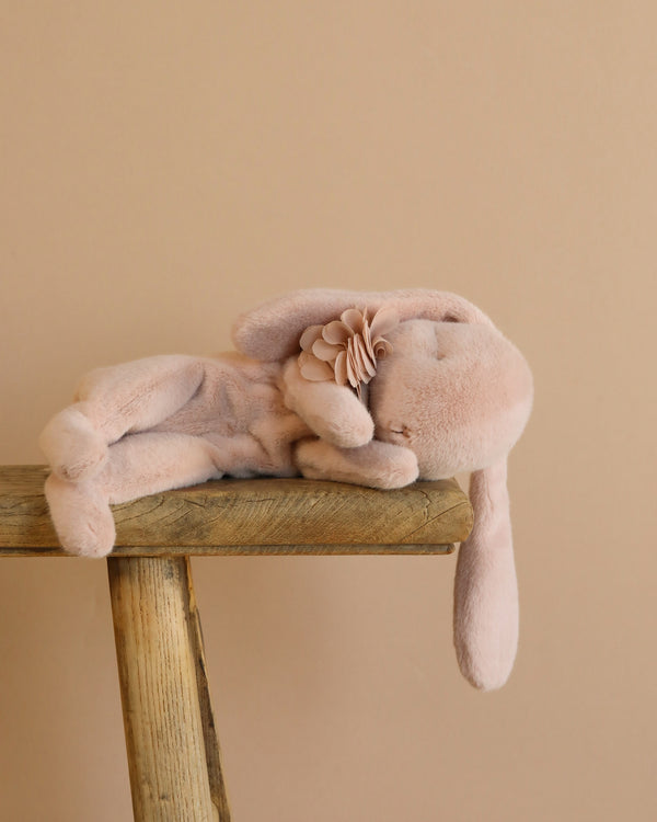 A Maileg Plush Sleeping Bunny - Powder, crafted from soft cotton fabric, reclines on its side on a wooden stool against a beige backdrop, gently holding a matching flower.