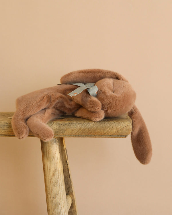 The Maileg Plush Sleeping Bunny - Soft Brown, a cotton rabbit with floppy ears, rests on its side like it’s sleeping. It balances on a wooden stool and wears a gray ribbon around its neck against a plain beige backdrop.
