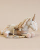 A Maileg Unicorn with a golden horn and multicolored yarn mane, made from recycled polyester, lies on a soft blanket against a light beige background.