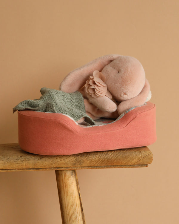 The Maileg Sleeping Bunny With Bed includes a plush bunny with a flower accessory, nestled in a cozy recycled polyester bed. This pink pet bed rests on a wooden stool, featuring a green knitted blanket against a soft beige backdrop.