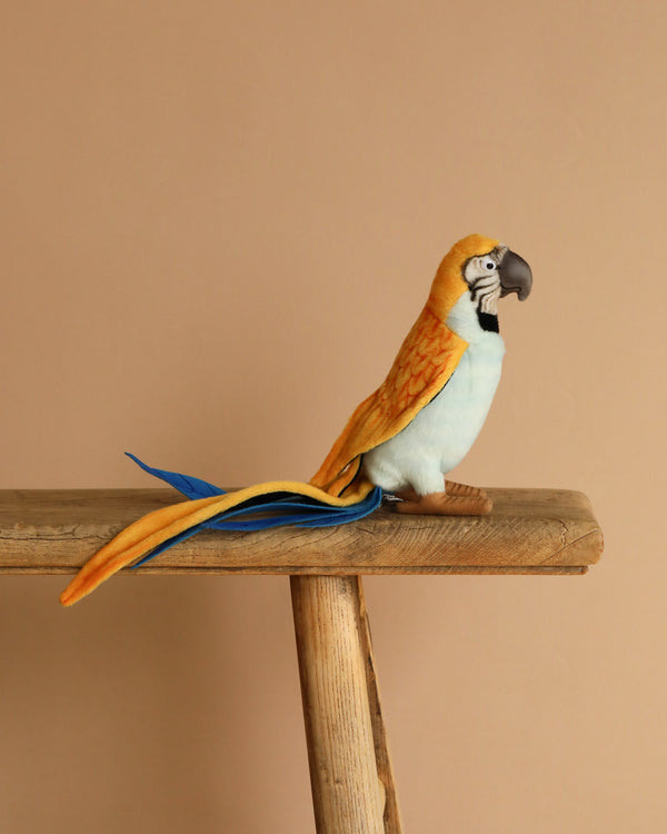 A Parrot Stuffed Animal, featuring artisanal craftsmanship and vibrant yellow, blue, and orange feathers, perches on a wooden beam against a beige background.
