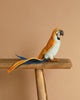 A Parrot Stuffed Animal, featuring artisanal craftsmanship and vibrant yellow, blue, and orange feathers, perches on a wooden beam against a beige background.