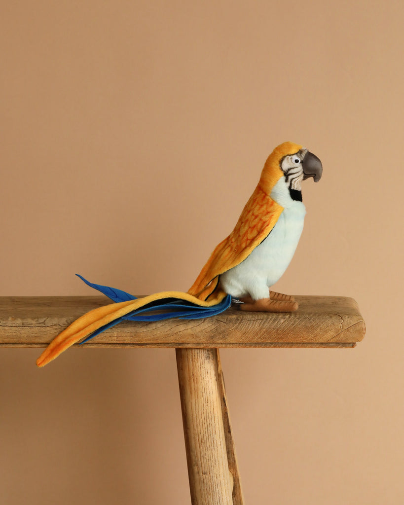 A Parrot Stuffed Animal, featuring artisanal craftsmanship and vibrant yellow, blue, and orange feathers, perches on a wooden beam against a beige background.