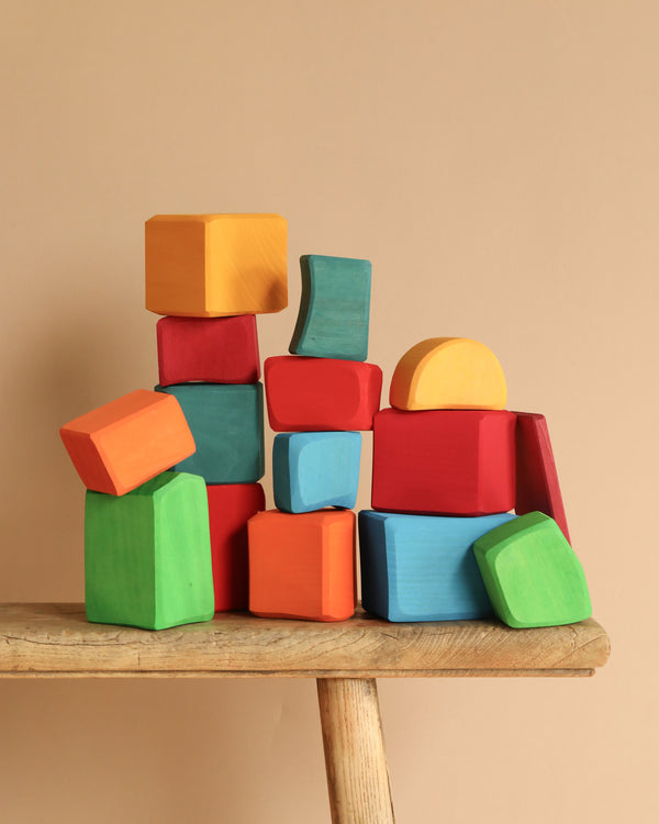 Grimm's Colored Waldorf Blocks, painted in vibrant orange, red, blue, green, and yellow hues, are stacked atop a wooden stool against a beige background. These non-toxic wooden blocks form a playful and balanced arrangement ideal for open-ended play.