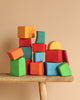 Grimm's Colored Waldorf Blocks, painted in vibrant orange, red, blue, green, and yellow hues, are stacked atop a wooden stool against a beige background. These non-toxic wooden blocks form a playful and balanced arrangement ideal for open-ended play.