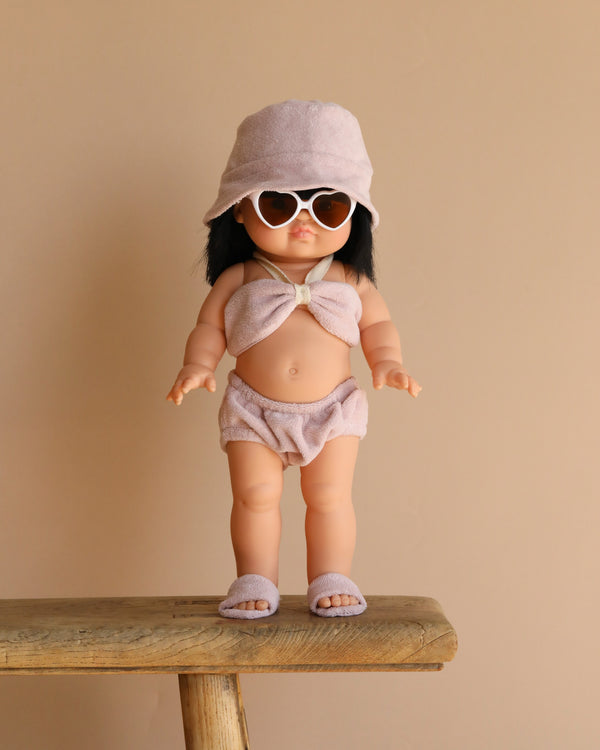 The Minikane Standing Doll (14") - Jade, exuding a pleasant vanilla scent, is dressed in a light pink two-piece outfit with heart-shaped sunglasses, a bucket hat, and sandals as it stands on a wooden bench against a plain beige background.