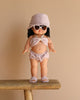 The Minikane Standing Doll (14") - Jade, exuding a pleasant vanilla scent, is dressed in a light pink two-piece outfit with heart-shaped sunglasses, a bucket hat, and sandals as it stands on a wooden bench against a plain beige background.