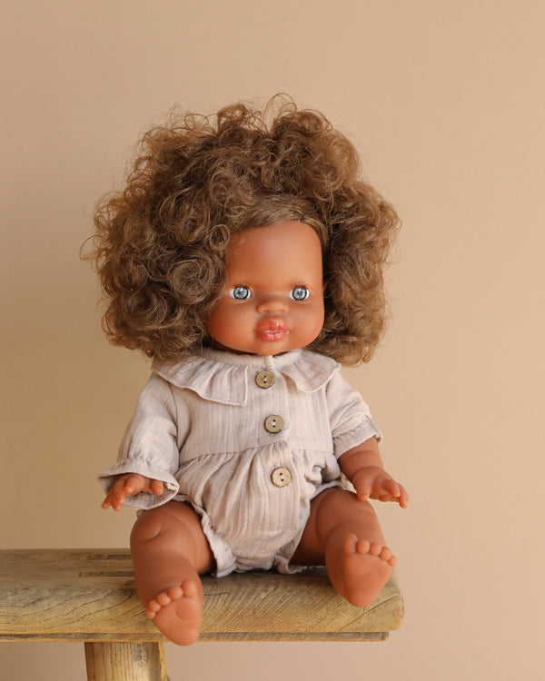 The Minikane Sitting Doll (13") - Charlie with curly brown hair wears a beige collared outfit with buttons, sitting on a wooden stool against a neutral background, and emits a gentle vanilla scent.