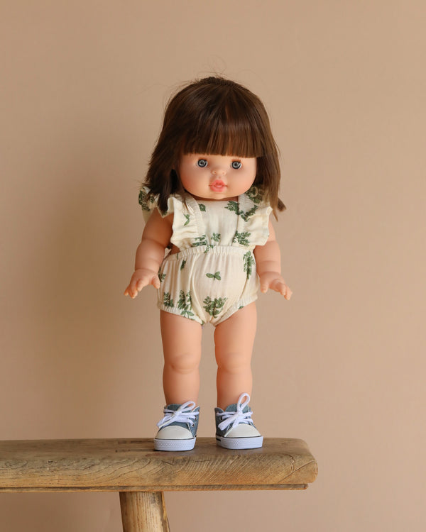 On a wooden bench against a beige background, the Minikane Standing Doll Chléa, featuring brown hair and wearing a floral outfit with blue sneakers, can be seen.