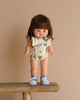 On a wooden bench against a beige background, the Minikane Standing Doll Chléa, featuring brown hair and wearing a floral outfit with blue sneakers, can be seen.