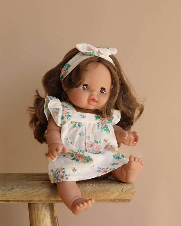 A Minikane Baby Doll, Leopoldine (13"), with long brown hair sits on a wooden stool in a white floral dress and matching headband. The doll exudes a natural vanilla scent against the plain light brown background.