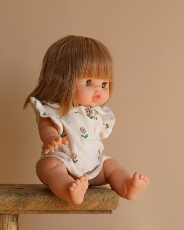 The Minikane Baby Doll (13") - Zoe is seated on a wooden ledge against a beige background. She has light brown hair and wears a white romper adorned with small flowers, her anatomically correct arms and legs slightly extended.
