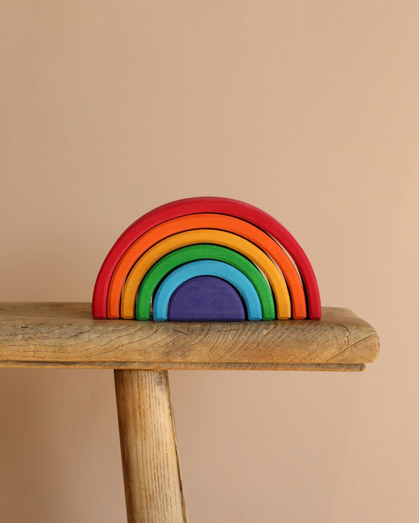 The Grimm's Wooden Rainbow, a non-toxic stacker toy with seven vibrant arcs from red to purple, is creatively arranged in a semicircle on a wooden bench against a beige background.