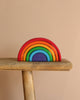 The Grimm's Wooden Rainbow, a non-toxic stacker toy with seven vibrant arcs from red to purple, is creatively arranged in a semicircle on a wooden bench against a beige background.