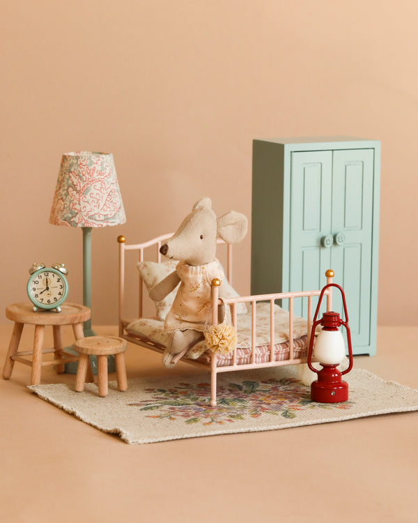 A Maileg Little Sister Bedtime Set features a stuffed toy mouse in a dress on a vintage bed, surrounded by charming mouse accessories including a rug, lamp, clock, two stools, red lantern, and pale blue wardrobe.